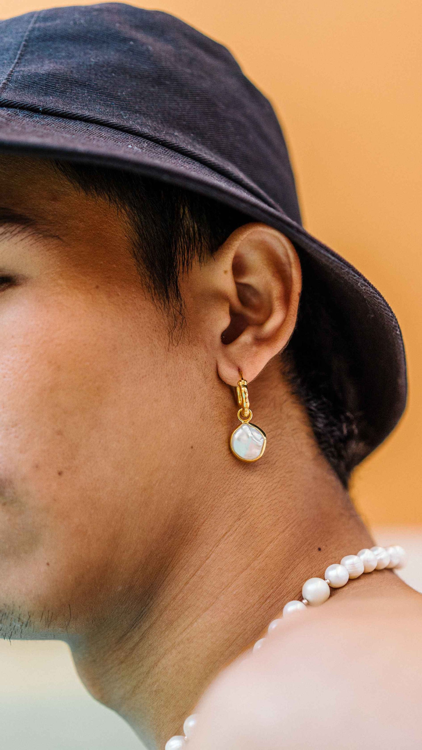 Close up of Male model in black bucket hat wearing the cereal pearl charm paired with chunky gold hoops. He is also wearing a Pearl Necklace mixed with South Se and Freshwater pearls.